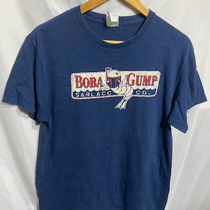 Könnte beinhalten: Marineblaues T-Shirt mit dem Aufdruck "Boba Gump Sarlacc Co." im Used-Look. Die Grafik zeigt eine Cartoon-Darstellung von Boba Fett und einem Sarlacc. Das T-Shirt hängt an einem Holzkleiderbügel.