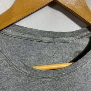 May include: Close-up of a gray t-shirt neckline with a wooden hanger. The shirt is made of a soft, textured fabric. The neckline is round and has a stitched hem. The wooden hanger is light brown.