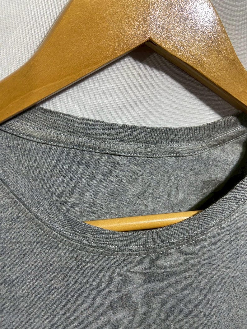 May include: Close-up of a gray t-shirt neckline with a wooden hanger. The shirt is made of a soft, textured fabric. The neckline is round and has a stitched hem. The wooden hanger is light brown.