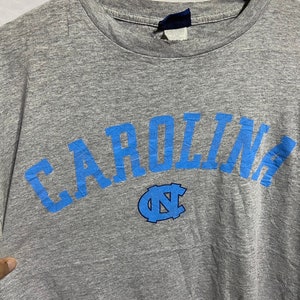 Puede incluir: Camiseta gris con un arco azul "CAROLINA" y un logotipo azul "NC".