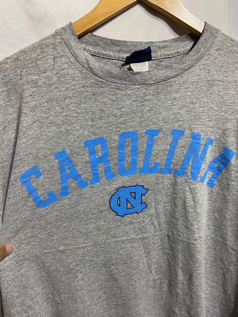 Puede incluir: Camiseta gris con un arco azul "CAROLINA" y un logotipo azul "NC".