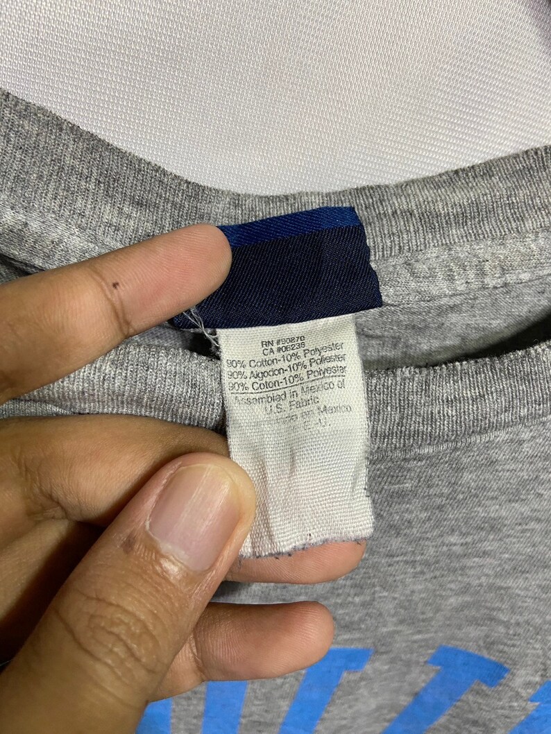 Puede incluir: Una camiseta gris con una etiqueta azul que dice "RN #90870 CA #06238 90% Algod&oacute;n-10% Poli&eacute;ster 30% Aigodon-10% Poliester 30% Algod&oacute;n-10% Poli&eacute;ster Ensamblado en M&eacute;xico de US. Tela Hecho en Mexico"