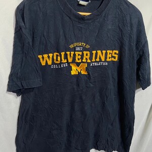 Może przedstawiać: Granatowa koszulka z napisem "PROPERTY OF WOLVERINES COLLEGE ATHLETICS" w złotym kolorze. Litera "M" jest umieszczona centralnie pod tekstem. Koszulka wisi na drewnianym wieszaku.