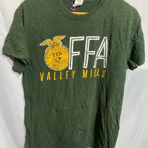 May include: Dark green t-shirt with the letters "FFA" in white and a gold emblem. Below the letters, the words "VALLEY MILLS" are printed in yellow. The shirt is hanging on a wooden hanger.