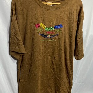 以下が含まれることがあります： ヤシの木のあるハンモックのシーンの上に「No Problem Mon Jamaica」と刺繍された茶色のTシャツ。 このシャツは半袖で、クルーネックです。