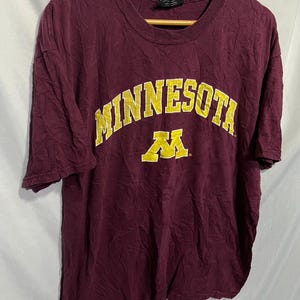 Könnte beinhalten: Ein maroonfarbenes T-Shirt mit einem goldenen "Minnesota" und "M" Logo. Das Shirt ist aus einem weichen, bequemen Material.