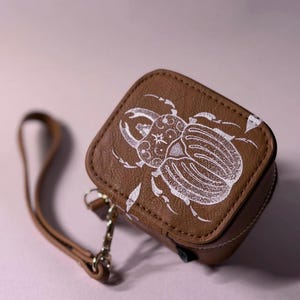 May include: A small, square, brown case with a textured surface featuring a white, detailed illustration of a beetle on the top lid. The case includes a matching brown wrist strap attached by a gold-toned metal clasp and hardware.