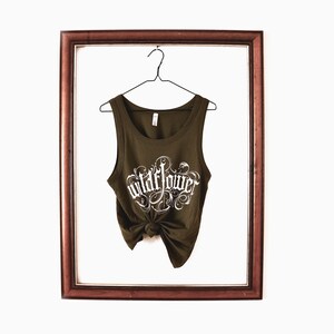 May include: An olive green sleeveless tank top with a scoop neckline, hanging on a wire hanger. The front features white decorative script text that reads wildflower, and the bottom hem is tied into a knot at the centre for a gathered look.