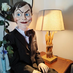 Puede incluir: Un muñeco de ventrílocuo sentado, con la cara pintada, vestido con un traje negro, camisa blanca y pajarita. El muñeco tiene ojos azules y una sonrisa siniestra. Una lámpara con base de madera y pantalla beige está detrás.