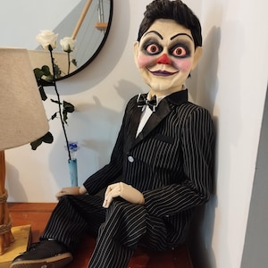 Peut inclure: Une poupée effrayante portant un costume rayé noir et blanc avec un nœud papillon noir. La poupée a un visage peint avec des lèvres rouges, un nez rouge et de grands yeux rouges. La poupée est assise sur une surface en bois.