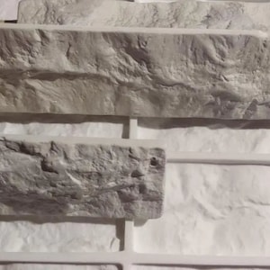 May include: Close-up of a white faux stone wall panel. The panel has a rough, textured surface that resembles natural stone. The panel is made of a lightweight material and is easy to install.