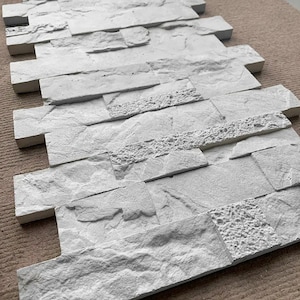 May include: White faux stone wall paneling with a textured surface. The paneling is made of a lightweight material and is easy to install.