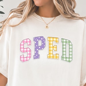 Peut inclure: Un t-shirt à manches courtes blanc cassé avec le texte SPED en grandes lettres style universitaire. Chaque lettre a un motif distinct : le S est à carreaux vichy roses, le P est violet à pois blancs, le E est à carreaux vichy jaunes et le D est à carreaux vichy verts.