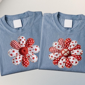 May include: Two pale blue t-shirts, each featuring a large appliqued flower. The flowers have red and white petals with polka dots, gingham, and maple leaf patterns. The flower centres are red.