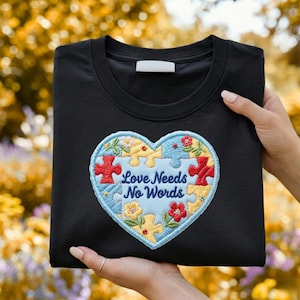 May include: Black crew neck sweatshirt with a heart-shaped patch. The patch features a puzzle design with the words "Love Needs No Words". The heart is decorated with flowers and puzzle pieces in red, yellow, and blue.