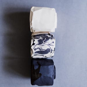 May include: Three square-shaped ceramic objects stacked vertically. The top object is solid matte white, the middle object features a black and white marbled pattern, and the bottom object is solid matte black. Each piece has a textured, uneven surface.