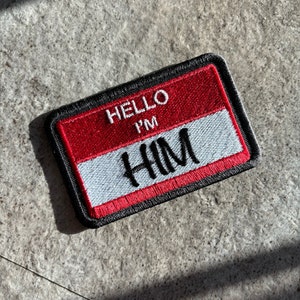 May include: A red and white name tag with black stitching. The tag says "Hello I'm HIM" in white embroidery.
