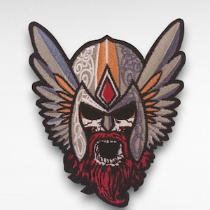 Peut inclure: Écusson brodé représentant un casque de Viking avec des ailes. Le patch présente un design détaillé avec une barbe rouge, une bouche ouverte et un casque avec des accents orange, gris et argent. Les ailes sont grises et bleues.