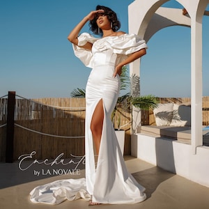 May include: A white satin off-the-shoulder wedding gown with a high slit and a flowing train. The dress features ruffled sleeves and a fitted bodice. The image is set against a clear blue sky and a white architectural backdrop.
