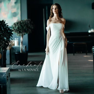 May include: A white off-the-shoulder wedding dress with a flowing skirt and a thigh-high slit. The dress is called "Enchanti by LA NOVALE".