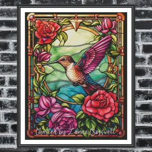 May include: Framed stained glass artwork depicting a hummingbird in flight, set amidst red and pink roses and green leaves. The background features blue and green hues. The piece is signed "Charted by Lanna Crosswell".