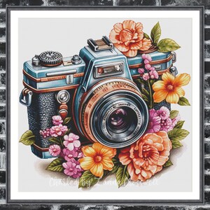 May include: A framed watercolour print featuring a vintage camera encircled by vibrant flowers. The camera is blue and black with a silver lens, decorated with orange, pink, and yellow blooms. The print is signed "Chasted by Lanna Crosswell".