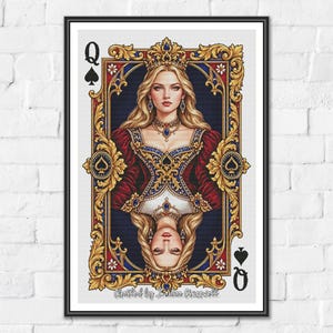 Op de afbeelding: Een ingelijste wandkunstprint die een Schoppenvrouw-speelkaart voorstelt. Het ontwerp toont een gespiegeld portret van een blond persoon met een kroon, een rood-blauwe jurk en sieraden, gevat in een sierlijke gouden lijst met schoppensymbolen.