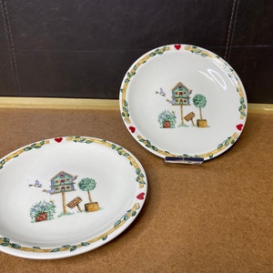 May include: Two white ceramic plates with a yellow and green leafy border and a red heart design. The plates feature a blue birdhouse with a red roof and a small green bush.