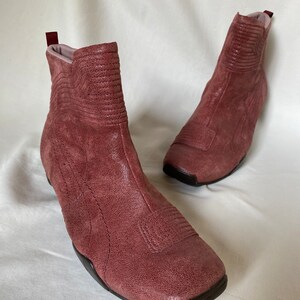 May include: A pair of dusty rose suede ankle boots with a square toe and a zipper on the back. The boots have a textured design and a black rubber sole.