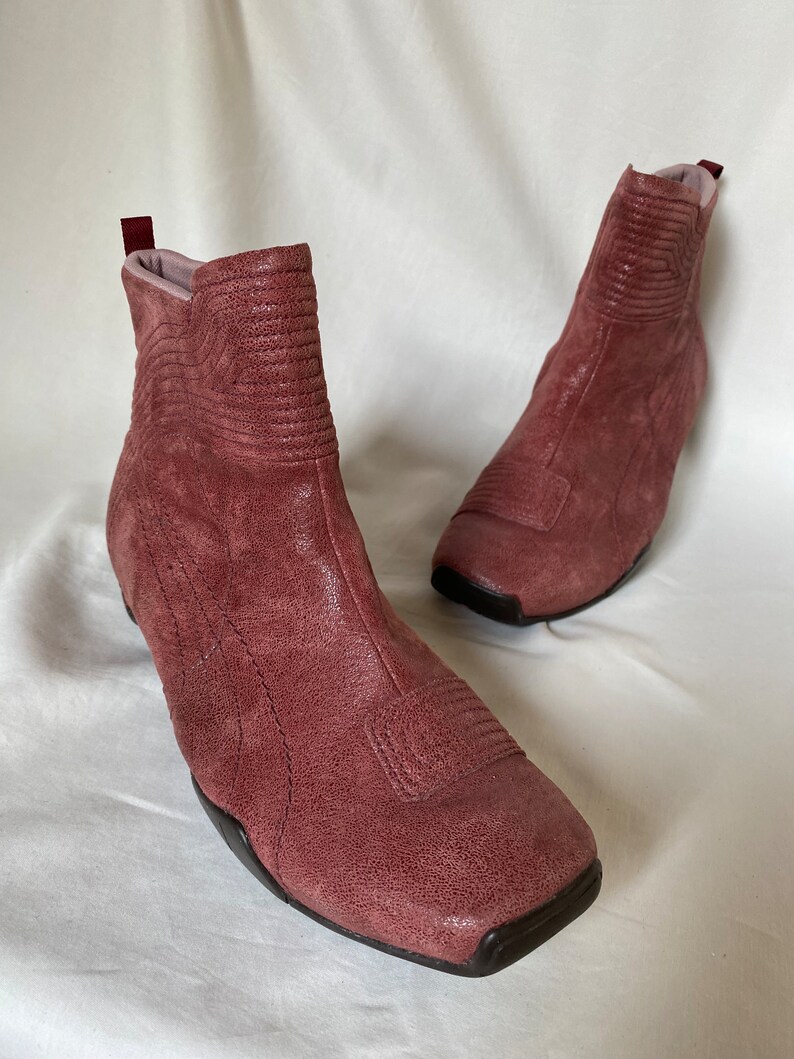 May include: A pair of dusty rose suede ankle boots with a square toe and a zipper on the back. The boots have a textured design and a black rubber sole.