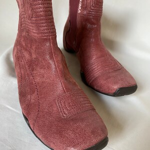 May include: A pair of dusty rose colored ankle boots with a square toe and a pull-on design. The boots have a textured leather upper and a black rubber sole.