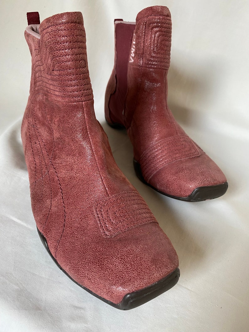 May include: A pair of dusty rose colored ankle boots with a square toe and a pull-on design. The boots have a textured leather upper and a black rubber sole.
