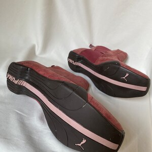 May include: A pair of burgundy suede Puma sneakers with pink accents. The shoes have a black rubber sole and a Puma logo on the side.