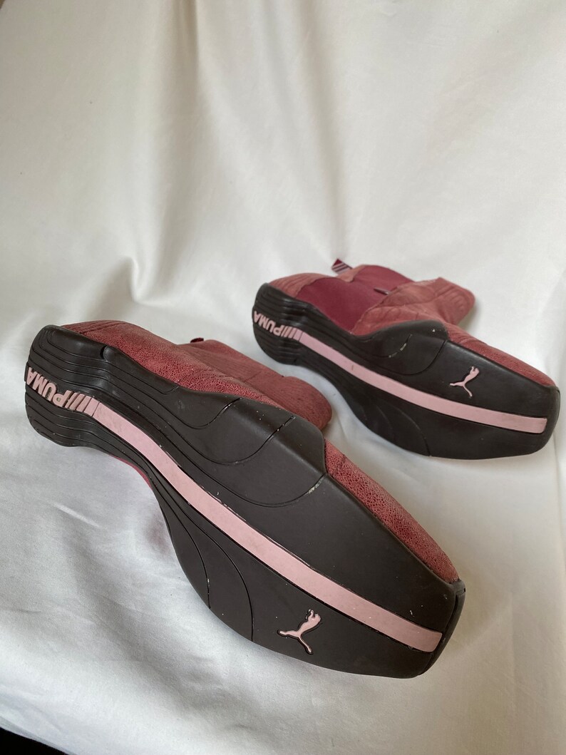 May include: A pair of burgundy suede Puma sneakers with pink accents. The shoes have a black rubber sole and a Puma logo on the side.