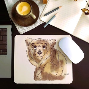 Può includere: Un tappetino per mouse beige con un'illustrazione di un orso bruno che guarda oltre la spalla. L'orso ha un naso marrone chiaro e occhi marroni scuri. Il testo "lau dar" è stampato nell'angolo in basso a destra del tappetino per mouse.