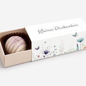 Può includere: Un tartufo al cioccolato bianco con striature viola in un pirottino di carta marrone, contenuto in una scatola rettangolare a scorrimento. L'esterno bianco presenta illustrazioni di fiori, farfalle e il testo tedesco Kleines Dankeschön.