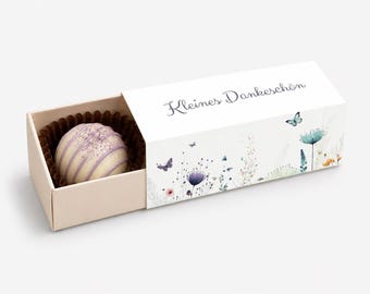 May include: A white chocolate truffle with purple drizzle in a brown paper liner, housed in a rectangular sliding box. The box features a white exterior with a floral and butterfly illustration and the German text Kleines Dankeschön.