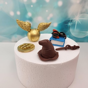 Könnte beinhalten: Ein weißer Kuchen, der mit einem goldenen Schnatz, einem braunen Zaubererhut, einem blauen Buch mit roten Brillen, einem goldenen 'H'-Symbol und einem braunen Zauberstab verziert ist.