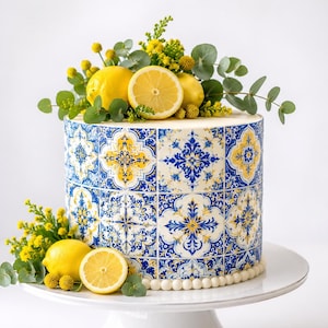 Op de afbeelding: Een ronde taart met een blauw, wit en geel tegelontwerp. De taart is versierd met verse citroenen, gele bloemen en groene bladeren. Hij staat op een witte taartstandaard.