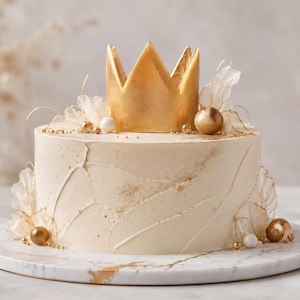 Könnte beinhalten: Ein runder Kuchen mit glattem, cremefarbenem Zuckerguss, strukturierten Pinselstrichen und goldenen Staubakzenten. Die Oberseite ist mit einer großen goldenen Krone, durchscheinenden weißen blütenblattartigen Verzierungen und Kugeln verziert.