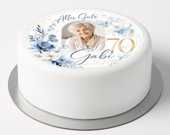 May include: A round white cake topper featuring a central photograph of a person, surrounded by blue and white floral illustrations, gold accents, and the text Alles Gute, 70, and Gabi in elegant blue and gold script lettering on a white background.
