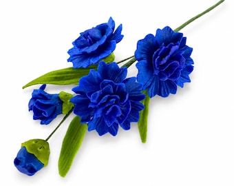 May include: A decorative floral spray featuring five vibrant blue carnation-style flowers in various stages of bloom, from a tight bud to fully opened blossoms, attached to thin green stems with several pointed green leaves extending outward.