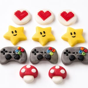 Könnte beinhalten: Neun bunte Fondantdekorationen im Videospiel-Thema. Die Dekorationen umfassen drei rote pixelige Herzen auf weißen Kreisen, drei gelbe lächelnde Sterne, drei graue Gamecontroller und zwei rot-weiße Pilze.