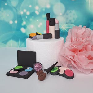 Könnte beinhalten: Eine Sammlung von Miniatur-Make-up-Artikeln, darunter Lippenstifte, Lidschattenpaletten und Pinsel, die auf einer weißen Plattform angeordnet sind. Die Lippenstifte sind in verschiedenen Farben erhältlich, und die Lidschatten umfassen Farbtöne von Rosa, Grün und Lila. Im Hintergrund befindet sich eine große rosa Papierblume.