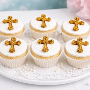 Può includere: Sei cupcake su un piatto bianco, ognuno sormontato da un disco bianco e una croce dorata. I cupcake hanno una base marrone chiaro e sono in pirottini di carta bianchi. Piccole perle bianche sono sparse.