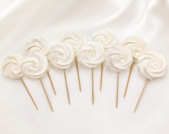 May include: A collection of ten white meringue toppers, each shaped like a swirling rose and mounted on a thin, pointed wooden stick. The meringues are arranged in a cluster, showcasing their smooth, spiraled texture and uniform off-white color.
