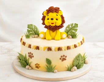 May include: A round cake with smooth cream-colored frosting, decorated with a yellow lion figurine on top. The sides feature brown paw prints, green fondant leaves, and grass accents. A border of small, alternating brown and yellow frosting dollops.