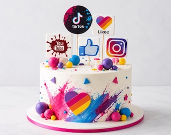 May include: A round white cake decorated with colorful paint splatters, small spheres, and a central heart shape. Topped with five social media themed signs featuring the logos for TikTok, YouTube, Likee, a thumbs-up icon, and Instagram on sticks.