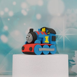 Könnte beinhalten: Ein blaues, rotes und gelbes Spielzeug-Zug, der an Thomas die Lokomotive erinnert, mit einem lächelnden Gesicht. Der Zug hat die Nummer "1" und ist vor einem verschwommenen, hellblauen Hintergrund platziert. Wahrscheinlich eine Tortenaufsatz.