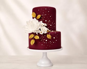 May include: A two-tier burgundy cake on a white pedestal stand. The cake is decorated with a large white flower, smaller white blossoms, and metallic gold leaves. Small white pearl-like accents are scattered across the smooth dark red surface.